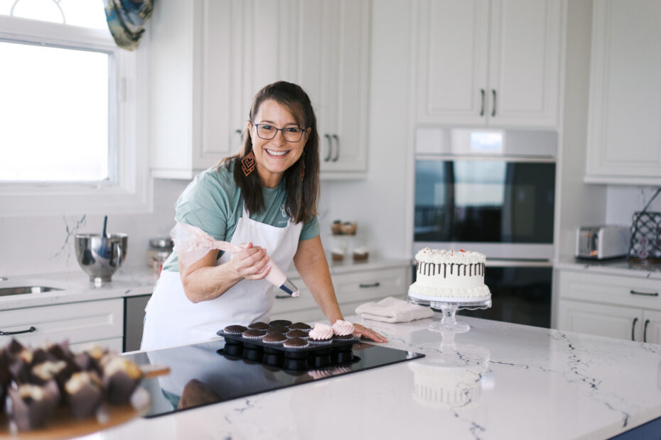 Brunette woman in a white apron ices cupcakes and smiles at the camera