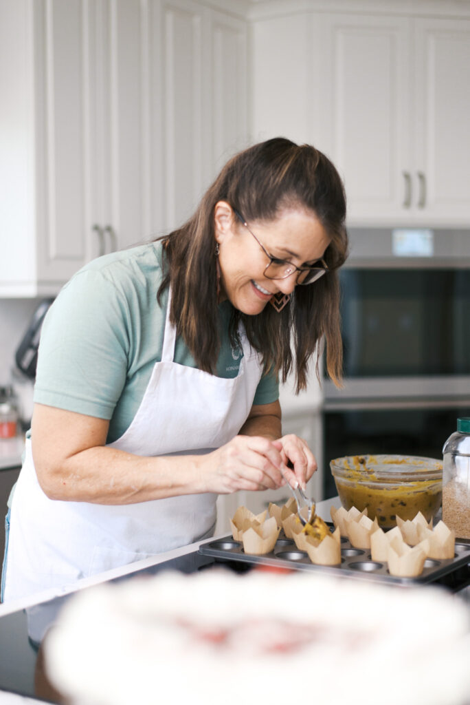 Brunette woman in a white apron spoons muffin batter into a muffin tin
