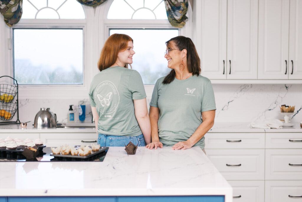 Two women, mother and daughter, stand in their home kitchen and smile at each other