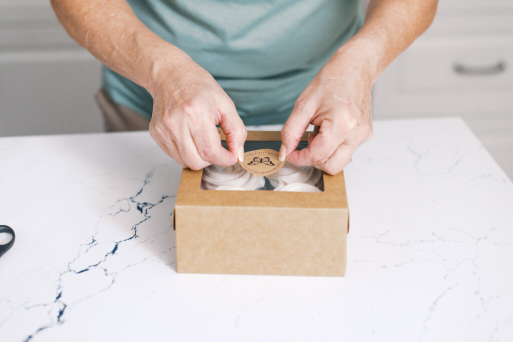 Baker applies a logo sticker to the top of a brown paper box that holds four cupcakes
