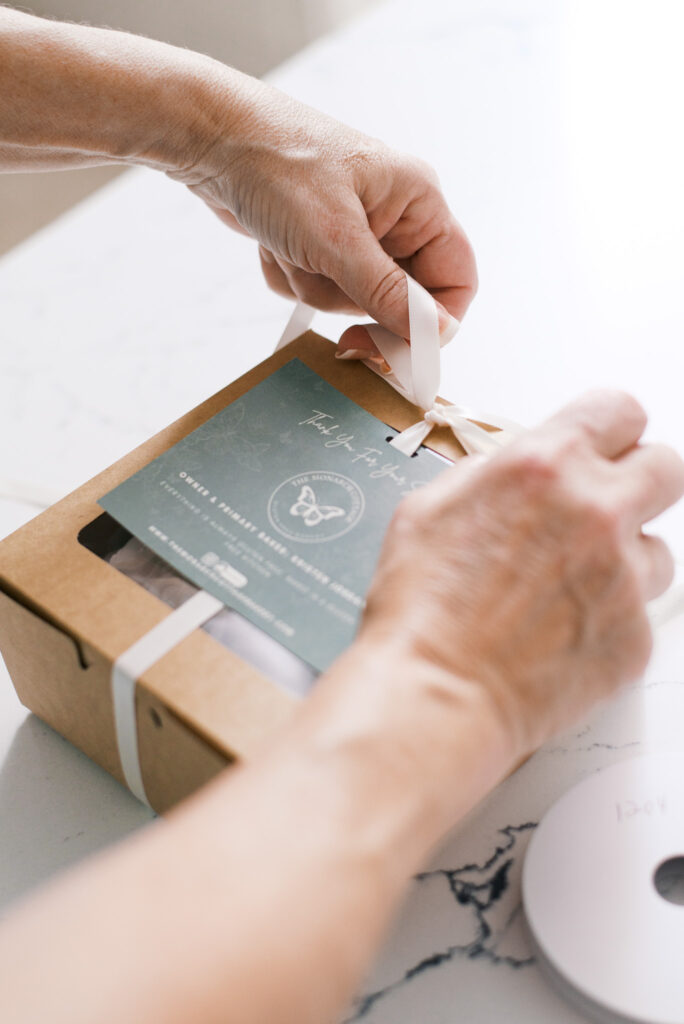 Baker ties a cream ribbon around a brown paper cupcake box, with a sage green thank you card on top