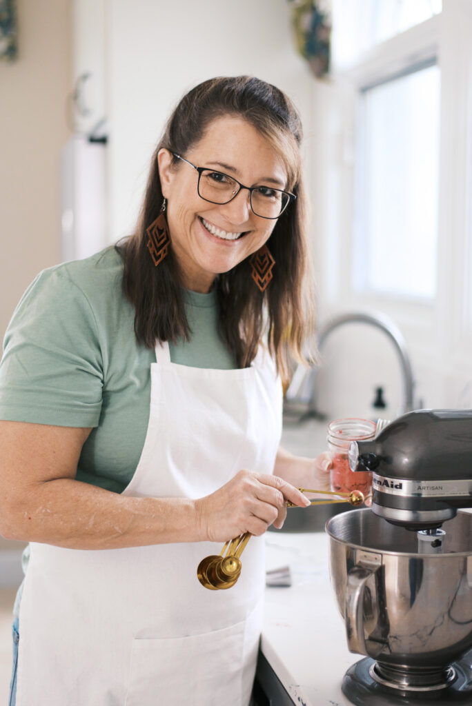 Brunette woman in a white apron measures baking ingredients into a mixer