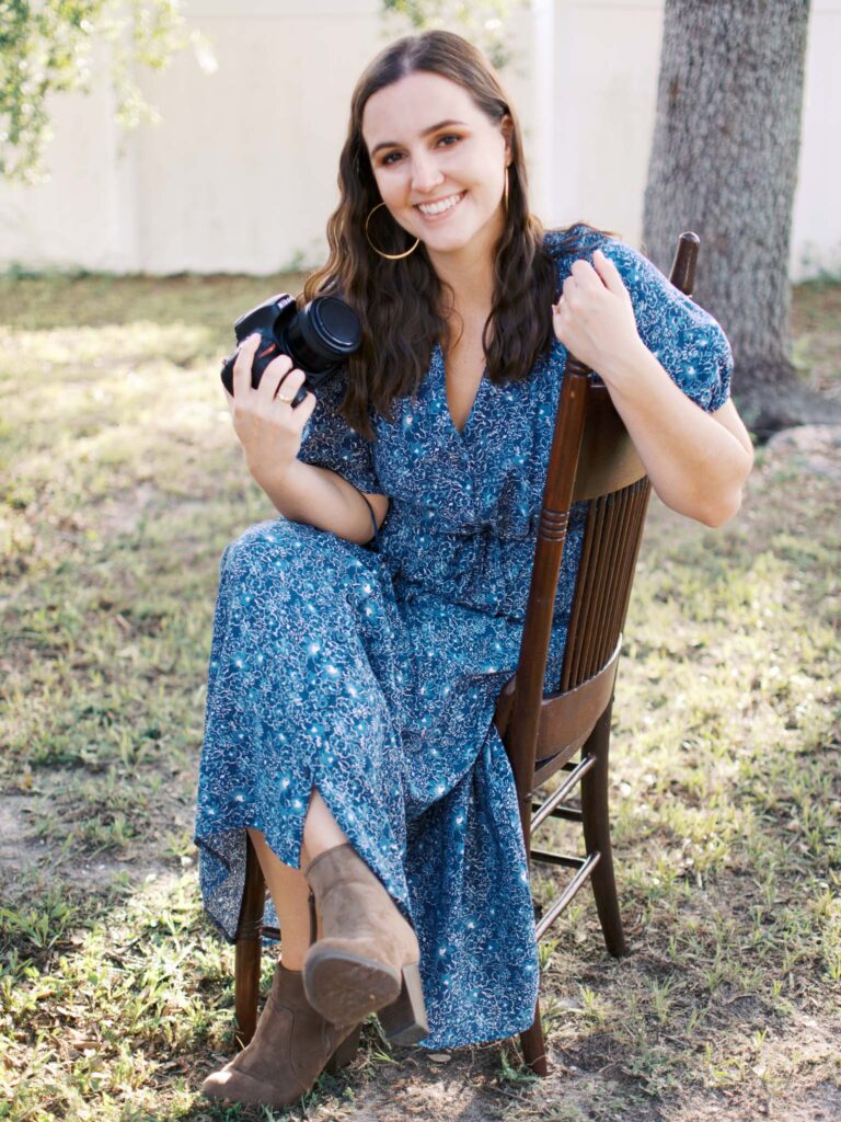 A brunette woman in a blue floral dress sits on a wooden chair outdoors, and smiles for the camera