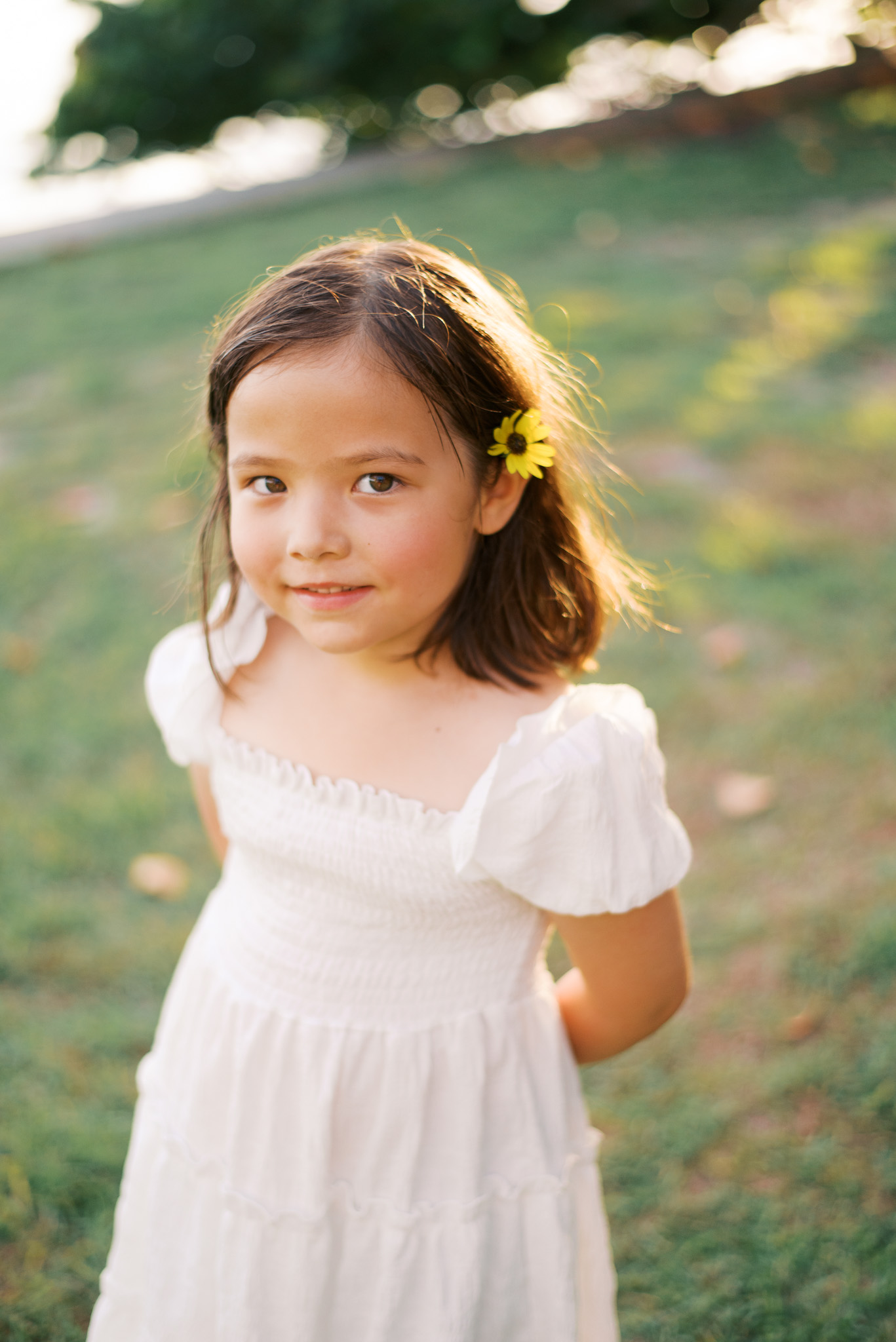 A young girl in a white dress with a wildflower in her hair smiles at the camera