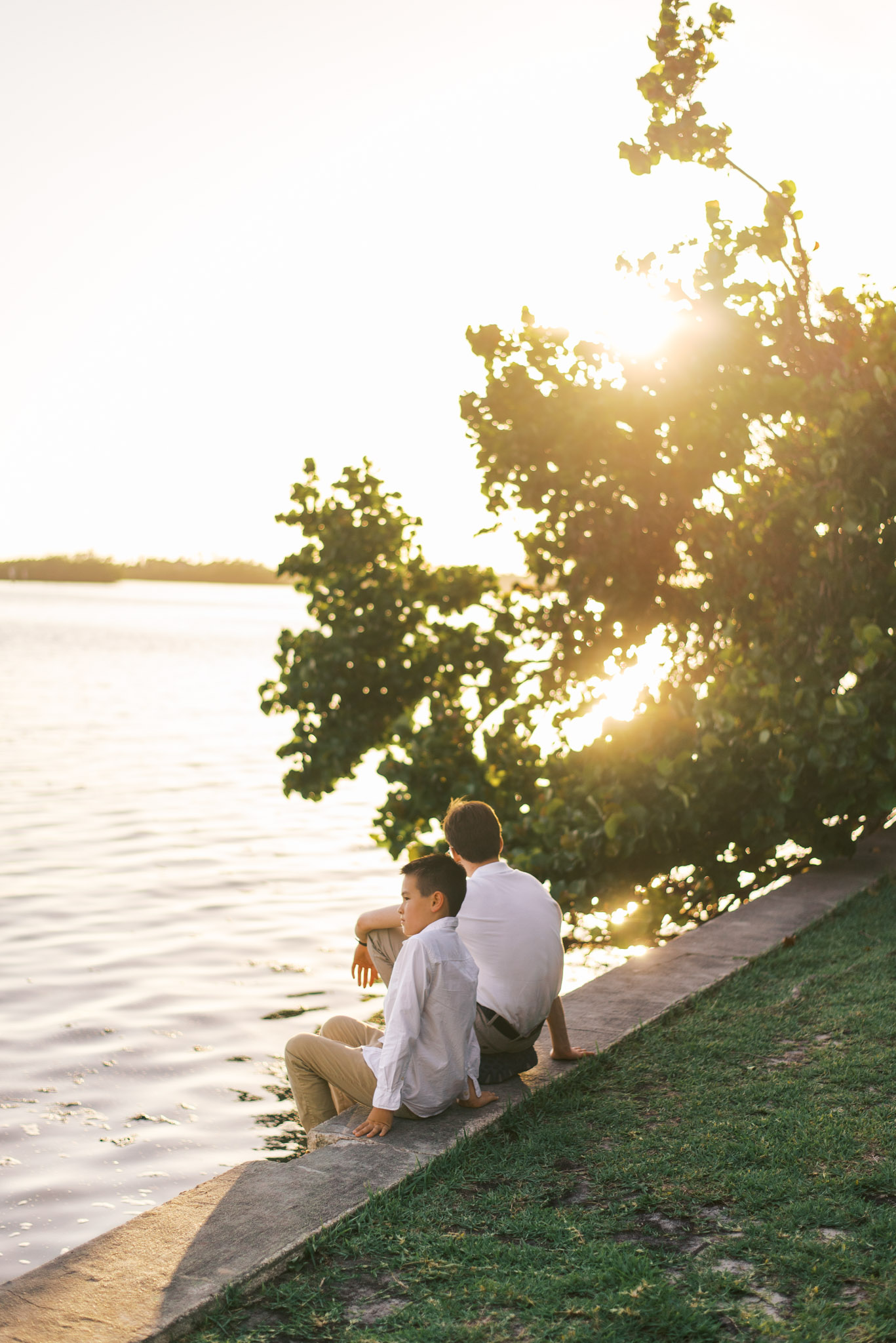 Two boys sit next to each other on a seawall, looking out over the water at the sunset