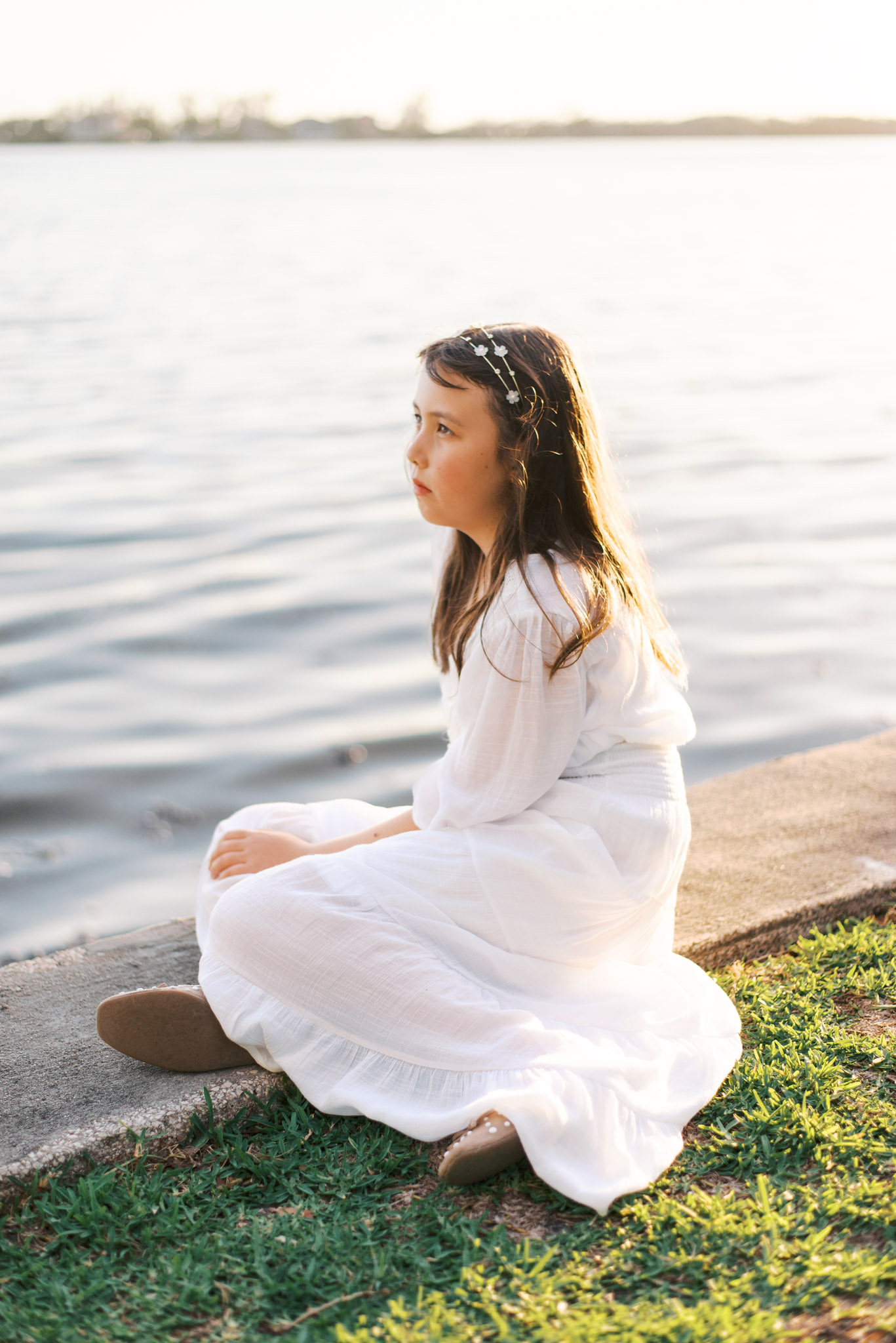 A young girl in a white dress sits in front of the water during sunset