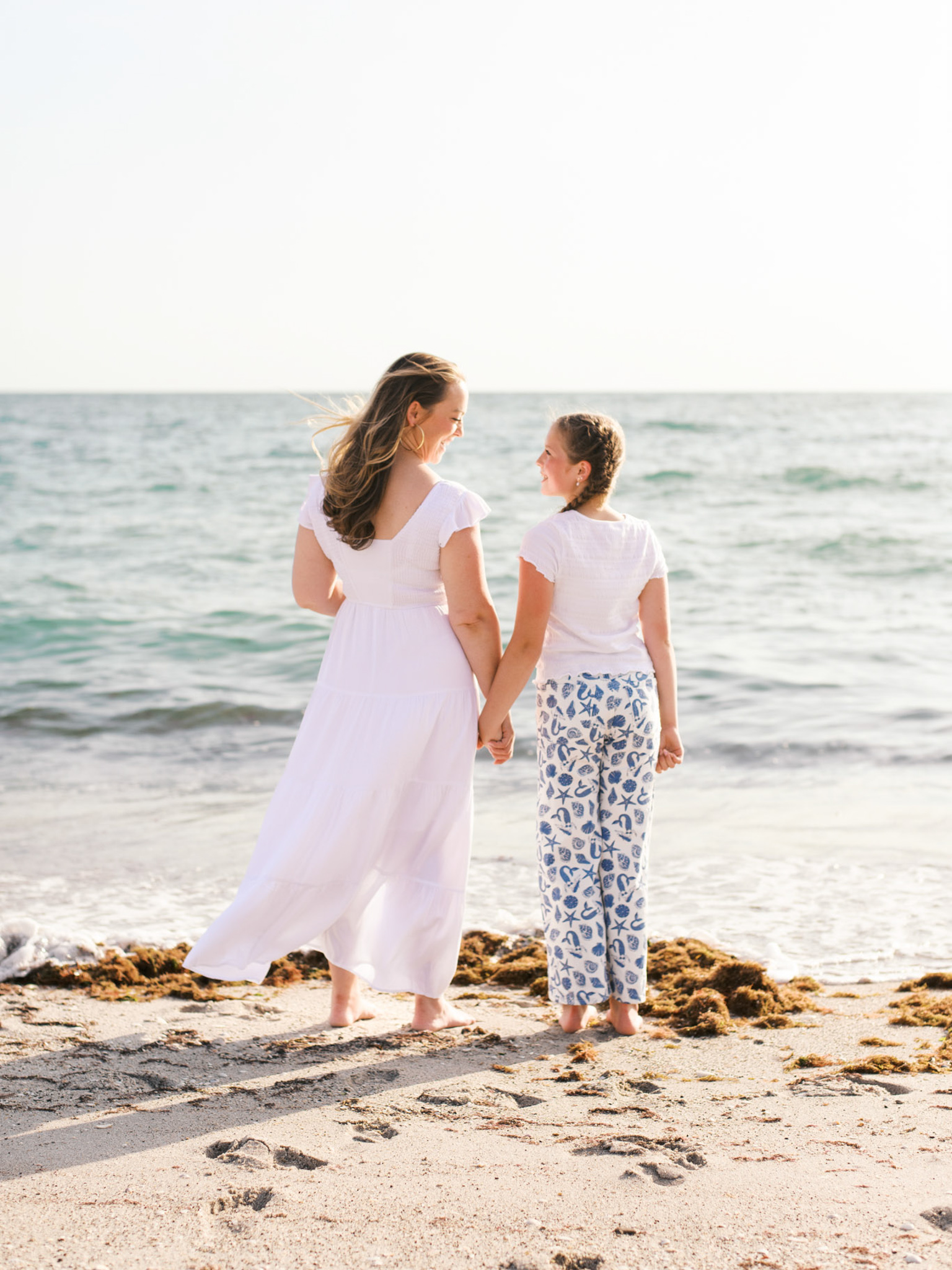 A woman and her young daughter stand on the beach facing the waves and look at each other