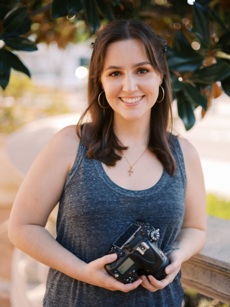 A brunette woman wearing a gray tank holds a camera in her hands and smiles