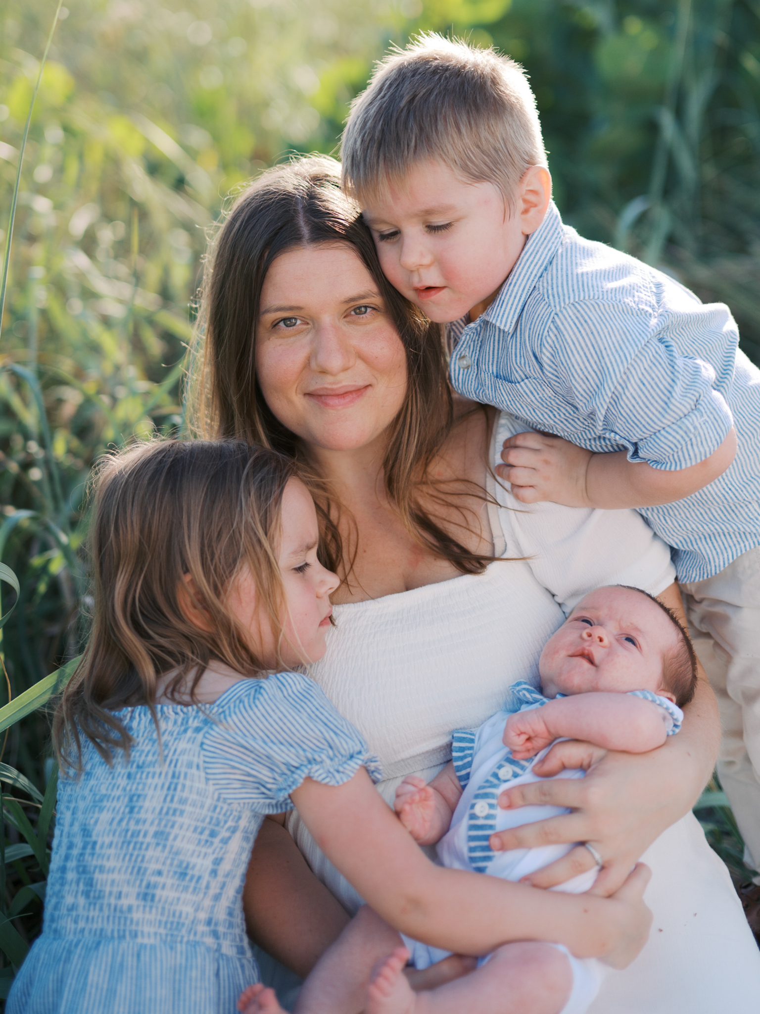 A young mother smiles, holding her newborn, while her young daughter and son hug her